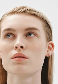 Pendientes de aro dorados con una pequeña gema roja, llevados en la oreja de un modelo. El modelo tiene piel clara y cabello rubio largo y liso.