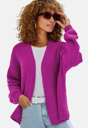 Fuchsiafarbener Strickcardigan mit offenem Vorderteil, gerippten Bündchen und legerer Passform. Kombiniert mit einem weißen Oberteil und hellen Jeans, dazu ein weißer Gürtel.