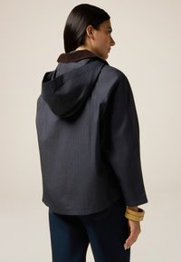 Veste bleu marine avec capuche, en tissu texturé, et col en velours côtelé marron. Coupe ample avec épaules tombantes, soulignant un design décontracté.