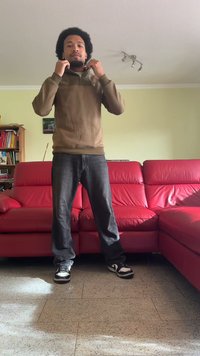 Ruda užtraukiama striukė su aukštu apykakle, tamsūs džinsai ir juodi bei balti sportbačiai, stovintis prieš raudoną odinį sofą.