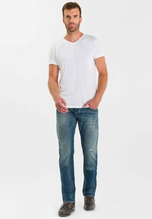 Uomo con T-shirt bianca casual a scollo a V, jeans blu e scarpe scure, in piedi davanti a uno sfondo bianco semplice.