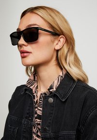 Ochelari de soare negri cu rame groase, textură netedă și lentile închise, purtați de o persoană cu păr lung, castaniu deschis și o cămașă cu un model.