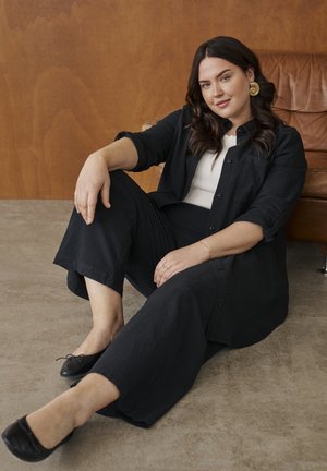 Femme aux cheveux foncés et boucles d'oreilles en or assise par terre, appuyée contre un fauteuil en cuir marron, portant un pantalon noir, une chemise noire, un top blanc et des ballerines noires.