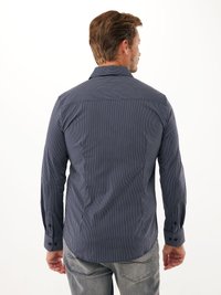 Uomo di spalle che indossa una camicia a maniche lunghe, blu navy con sottili strisce verticali bianche e jeans grigio chiaro su uno sfondo bianco.