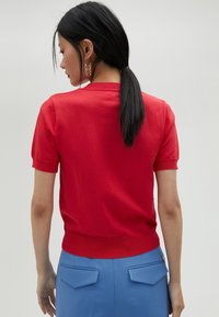 Pull court à manches courtes rouge avec des poignets et un ourlet côtelés, texture lisse. Assorti avec un pantalon bleu comportant deux poches à l'arrière.
