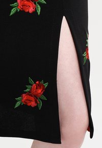 Czarny materiał z czerwonymi haftowanymi różami i zielonymi liśćmi, z pionowym rozcięciem odsłaniającym nagą skórę poniżej.