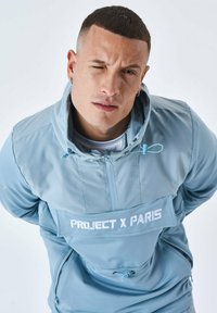 Ljusblå tröja med hög krage, framsida med dragkedja och stor patch som läser "PROJECT X PARIS." Den har elastiska ärmslut och dragsko.