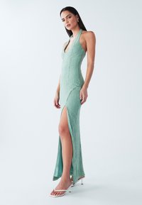 Robe dos nu vert menthe en tissu texturé, coupe ajustée, fendue à la cuisse. Associée à des sandales à talons blanches à brides.