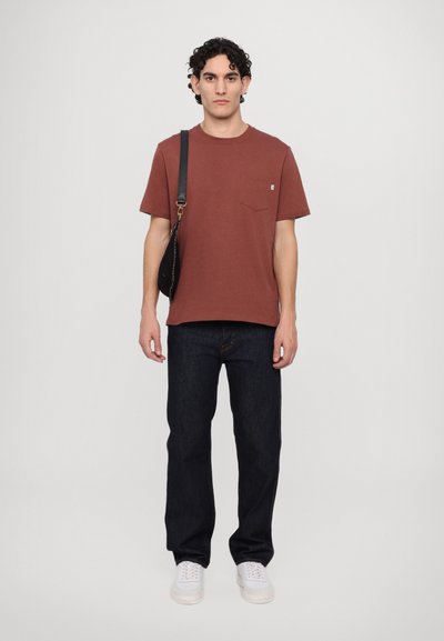 T-shirt couleur rouille avec une poche avant, associé à un jean bleu foncé et des baskets blanches. La tenue présente un design simple et une coupe décontractée.