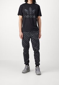 Svart t-shirt med stort mörkgrått Adidas-logotyp; matchande svarta joggers med upprepande mörkgrå logotypmönster; grå sportskor.