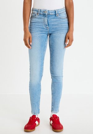 Persoon die lichtblauwe skinny jeans, witte sokken en rode veterschoenen draagt, staand tegen een effen witte achtergrond.