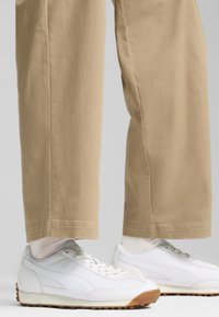 White sneakers with textured overlays and gum rubber soles, paired with loose beige trousers, displaying a casual, sporty look.