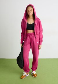 Um hoodie rosa com fecho éclair e um par de calças de moletom a condizer, sobre uma t-shirt cropped preta. Ténis atléticos castanhos e brancos, segurando uma bolsa preta.