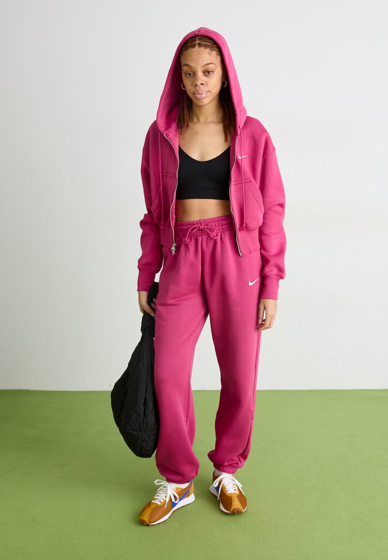 Różowa bluza na zamek z pasującymi dresami, założona na czarny crop top. Brązowe i białe buty sportowe, trzymająca czarną torbę.