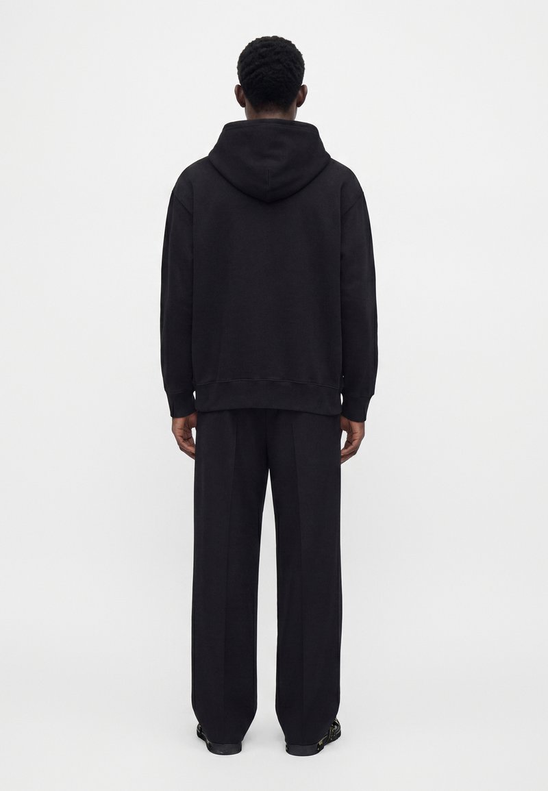 Homme debout de dos, portant un sweat à capuche noir et un pantalon noir ample sur un fond blanc uni.