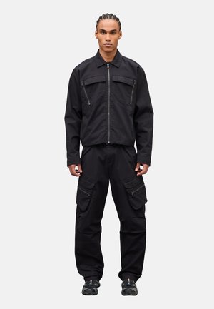 Hombre con cabello trenzado de pie, vistiendo una chaqueta negra con cremallera y solapas, con bolsillos con cremallera y botones, pantalones cargo a juego y zapatillas negras, sobre un fondo blanco.