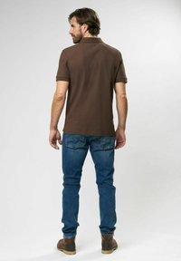 Braunes Poloshirt mit kurzen Ärmeln, kombiniert mit blauen Jeans. Das Shirt hat einen klassischen Kragen und eine glatte Textur. Stiefel vervollständigen das Outfit.