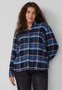 Woman with long curly hair wearing a blue and black plaid button-up shirt and black pants, standing against a light gray background.