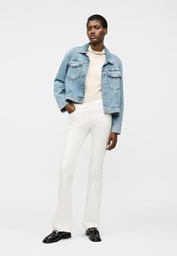 Γυναίκα που φοράει ανοιχτό μπλε τζιν μπουφάν, μπεζ μπλούζα, λευκό καμπάνα παντελόνι και μαύρα μοκασίνια, στέκεται με το ένα χέρι στην τσέπη σε λευκό φόντο.