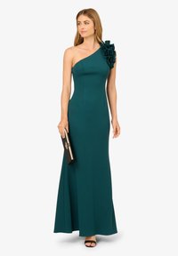 Tyrkysové šaty na jedno rameno s volánem na rameni; přiléhavý korzet, splývavá sukně; nošeno s černou kabelkou a sandály na otevřenou špičku.