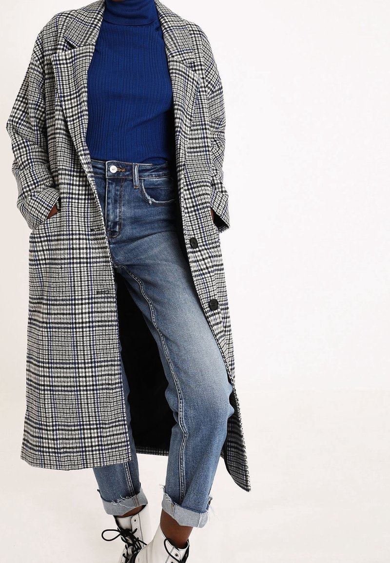 Cappotto lungo a scacchi bianco e nero con tasche laterali, indossato sopra un dolcevita blu a coste e jeans azzurri a risvolto, abbinato a stivali bianchi.