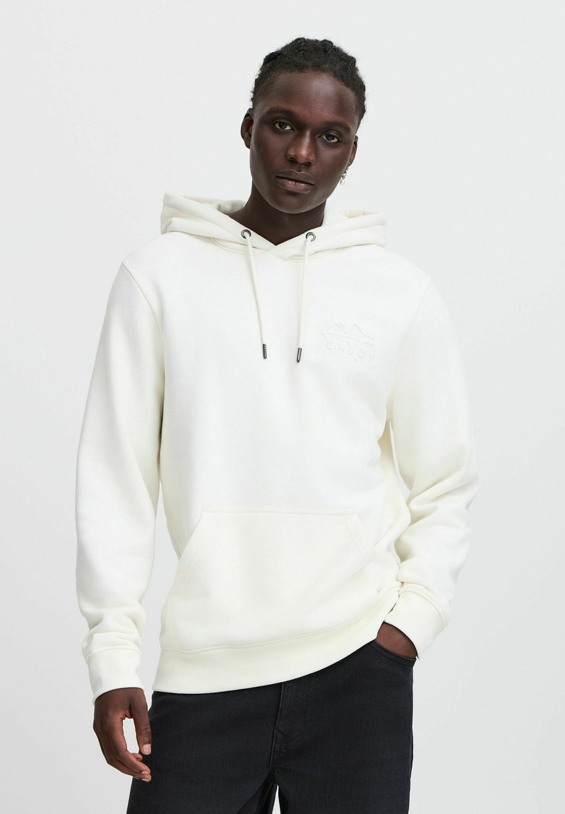 Witte hoodie van zacht materiaal, met een voorzak en trekkoorden. Subtiele "ENJOY" borduursel op de borst. Gepaarde met donkere spijkerbroek.
