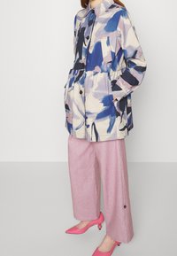 Nainen, joka pukeutuu siniseen, vaaleanpunaiseen ja valkoiseen abstraktiin kuvioituun takkiin, vaaleanpunaisiin leveälahkeisiin housuihin ja vaaleanpunaisiin teräväkärkisiin korkokenkiin, seisoo tasaisella taustalla.