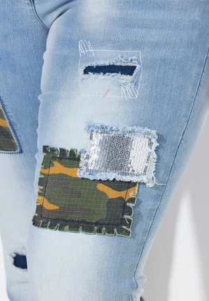 Blauwe denim jeans met meerdere patches: een camouflagepatch, een paillettenpatch en rafelige randen. Verschillende texturen en stikdetails zichtbaar.