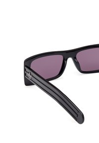 Ochelari de soare negri cu un design elegant și unghiular, având lentile de culoare purpurie închisă și brațe stripe negre. Include un detaliu mic cu logo pe braț.