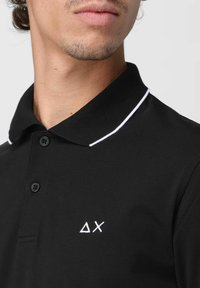 Polo nero con un accento bianco al colletto, dotato di tre bottoni neri e un piccolo logo bianco nell'area del petto sinistro.