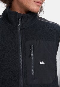 Black fleece vest with a high collar, front zipper, and a patch pocket. Features textured material and contrasting fabric accents.