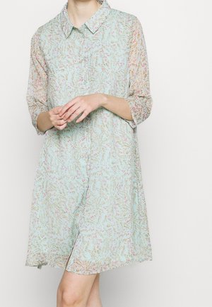 Femme portant une robe chemise bleu clair à fleurs, manches trois-quarts, col et ourlet à hauteur du genou, debout devant un fond blanc.