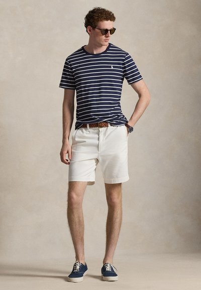 Άνδρας που φορά γυαλιά ηλίου, μπλουζάκι με μπλε-άσπρες ρίγες, λευκά σορτς με καφέ ζώνη, μπλε αθλητικά παπούτσια, στέκεται με το ένα χέρι στην τσέπη.