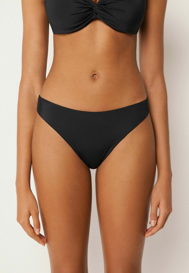 Femme portant un bas de bikini noir et un haut de bikini noir avec un détail torsadé, montrée du milieu de la poitrine au haut de la cuisse, debout.