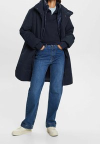 Abrigo acolchado azul marino sobre una camiseta oscura, combinado con jeans de tiro alto y zapatillas color crema. El abrigo tiene un cuello alto y bolsillos delanteros.