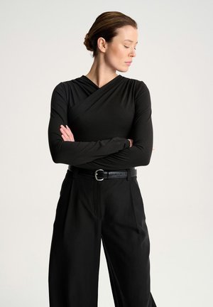 Kobieta z brązowymi włosami związanymi w koka, ubrana w czarną bluzkę z długim rękawem i czarne spodnie, stojąca z założonymi rękami, patrząca w lewo.