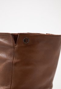 Botte en cuir marron avec une texture lisse et une légère brillance. Elle présente un accessoire en métal noir simple sur le côté, près de l'ouverture.