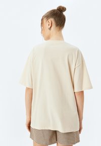 Camiseta de algodón beige oversize con mangas cortas y escote redondo, combinada con pantalones cortos a cuadros. Diseño sencillo, sin logotipos visibles.