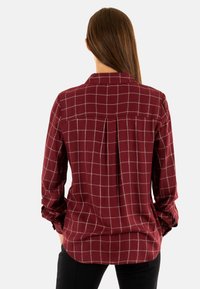 Camicia button-up bordeaux con motivo a quadri, maniche lunghe e dettaglio con piega sul retro. Texture liscia, colletto e accenti sui polsini.