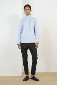 Světle modrý dlouhý rukáv s vysokým límcem, předním zapínáním, ve spojení s úzkými černými kalhotami a černými slip-on botami. Hladká textura.