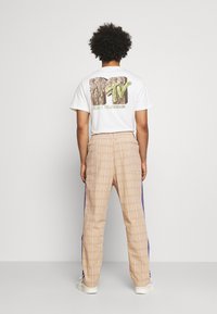 Valkoinen T-paita, jossa on monivärinen MTV-logo selässä, yhdistettynä vaaleanbeigeihin ruudullisiin housuihin, joissa on purppurat sivujuovat, ja valkoisiin lenkkareihin.