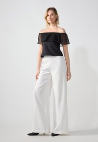 Top negru off-the-shoulder cu detalii transparente din volane, asortat cu pantaloni albi cu picior larg și pantofi negri cu vârful ascuțit. Texturi fine.