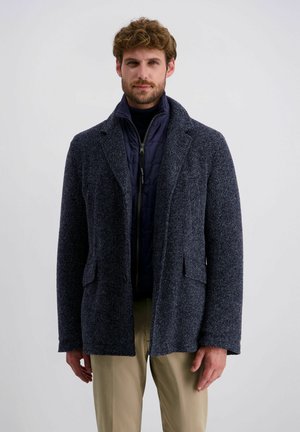 Mann mit lockigem Haar, der eine marineblaue gesteppte Jacke unter einem dunkelgrauen strukturierten Mantel und beige Hosen vor einem einfarbigen Hintergrund trägt.
