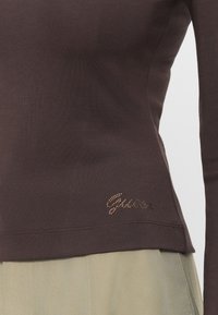 Top lung cu mânecă lungă, de culoare brună, cu un design ajustat, realizat dintr-un material neted. Prezintă un accent glossy cu logo din pietre strălucitoare la nivelul șoldului.