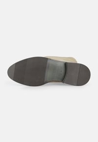 Leder-Schuh mit einem beigen Obermaterial und einer schwarzen Gummisohle, die horizontale Rillen für Traktion aufweist. Gestickte Akzente in der Nähe der Ferse.