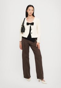 Ivory cropped jacket with black buttons and bow detail, black top, and brown pinstriped trousers. White pointed heels and black bag.