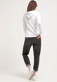 Sudadera blanca de algodón con capucha ajustable, combinada con jeans de corte relajado en gris oscuro. Zapatillas blancas completan el atuendo.