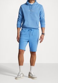 Man i ljusblå hoodie och matchande shorts med snörning, iförda vita sneakers och strumpor, stående mot en neutral bakgrund.