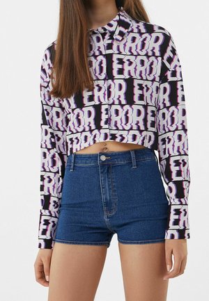 Vrouw draagt een cropped shirt met lange mouwen met herhalende "ERROR" tekst en een high-waisted blauwe denim short.