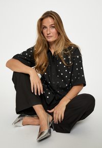 Čierna košeľa na gombíky s kovovými striebornými bodkami, skombinovaná s voľnými čiernymi nohavicami a striebornými špicatými topánkami.